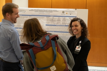 Wellbeing Fellow Gretchen Roman, DPT, PhD, discusses her project with Symposium attendees, a smiling man and a woman with her back to the camera.
