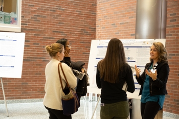 A woman discusses her Wellbeing Fellowship project in front of a poster with symposium attendees looking on