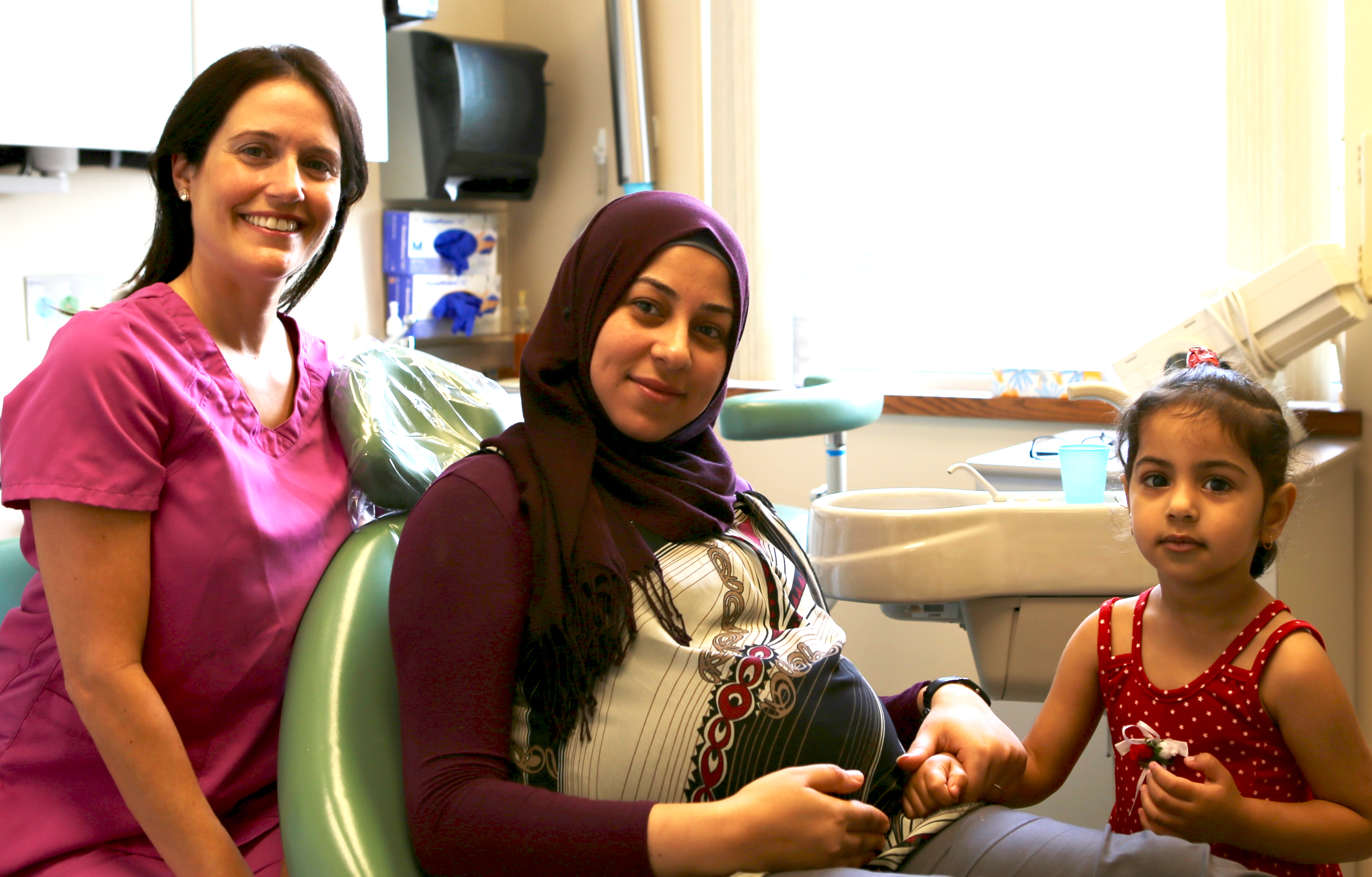 a pregnant woman sits in a dental chair, holding the hand of a child. A person in scrubs smiles next to them.