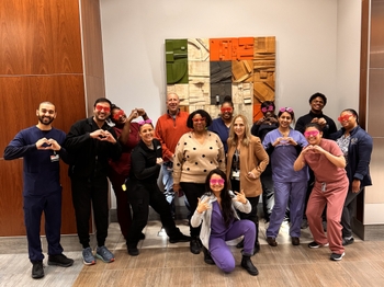 A group of people pose together in front of an abstract art piece. Most are wearing heart-shaped glasses or holding their hands up in heart shapes.