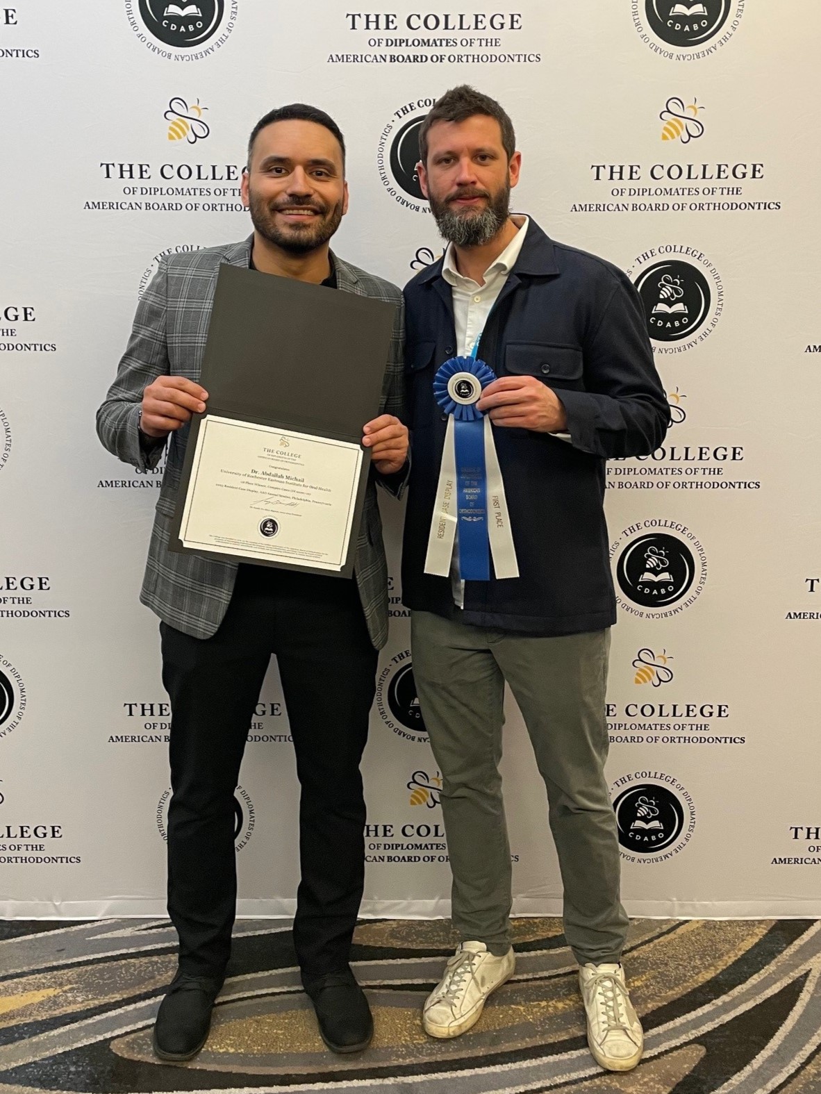Two men in suits stand in front of a backdrop reading "The College of Diplomats of the American Board of Orthodontics". One holds a certificate with the name "Dr. Abdallah Michail" and the other holds a blue and white ribbon that says "Resident Case Display First Place"