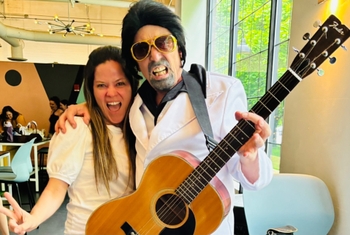 A woman makes a peace sign next to a man in a wig and sunglasses who is playing guitar.