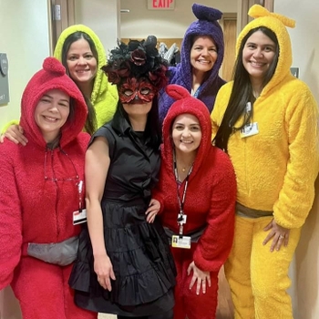 5 people dressed as teletubbies stand around a girl in black wearing a flowery mask.