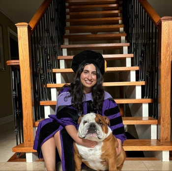 A woman in a purple graduation gown and cap sits on the stairs with an English bulldog.