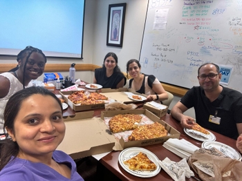 A group of people sit at a table in a break room sharing pizza.