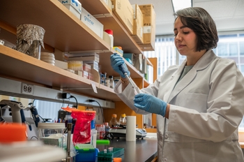 A woman in a lab coat stands at a lab bench holding a pipette.