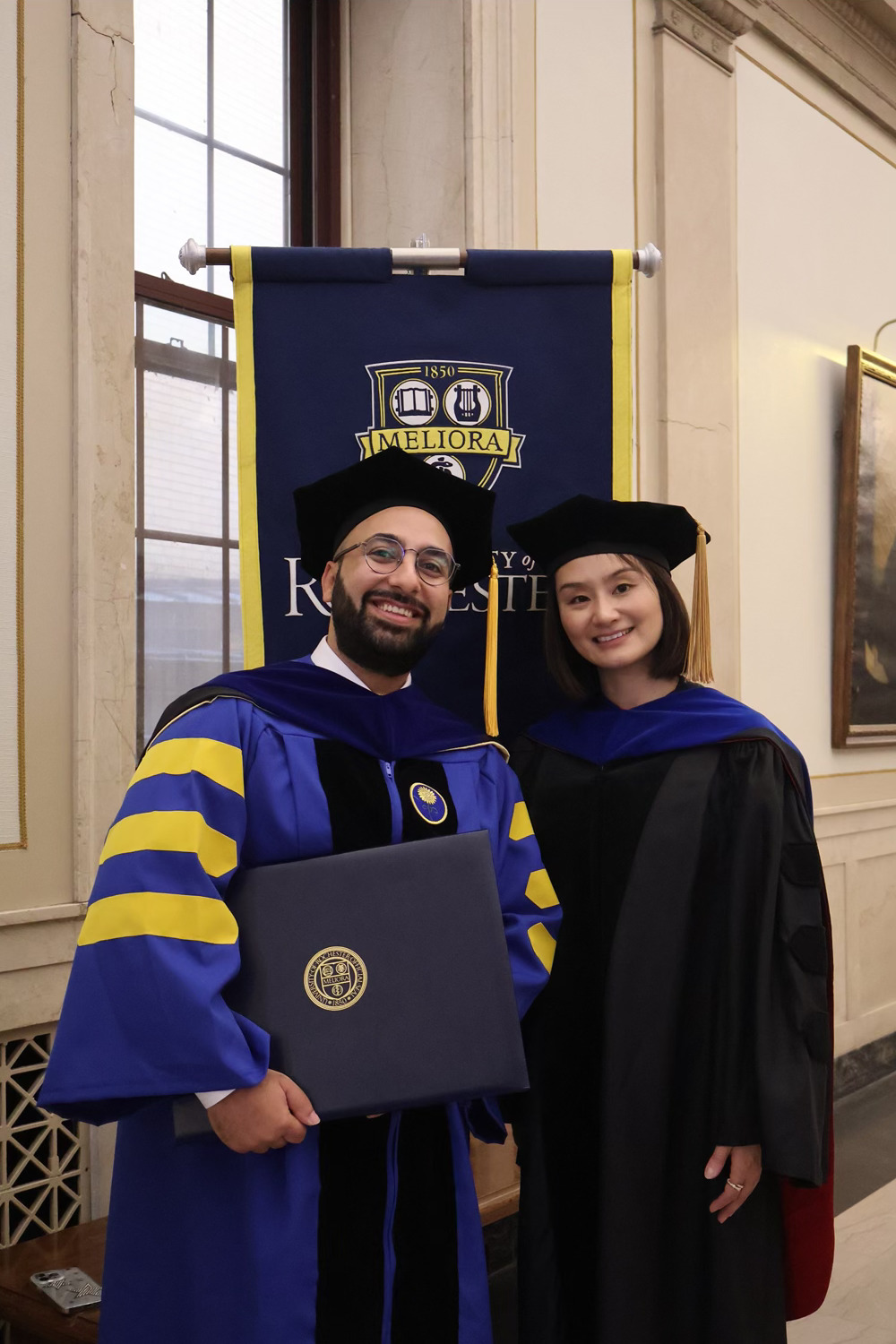 Two people in doctoral robes stand in front of a University of Rochester banner.
