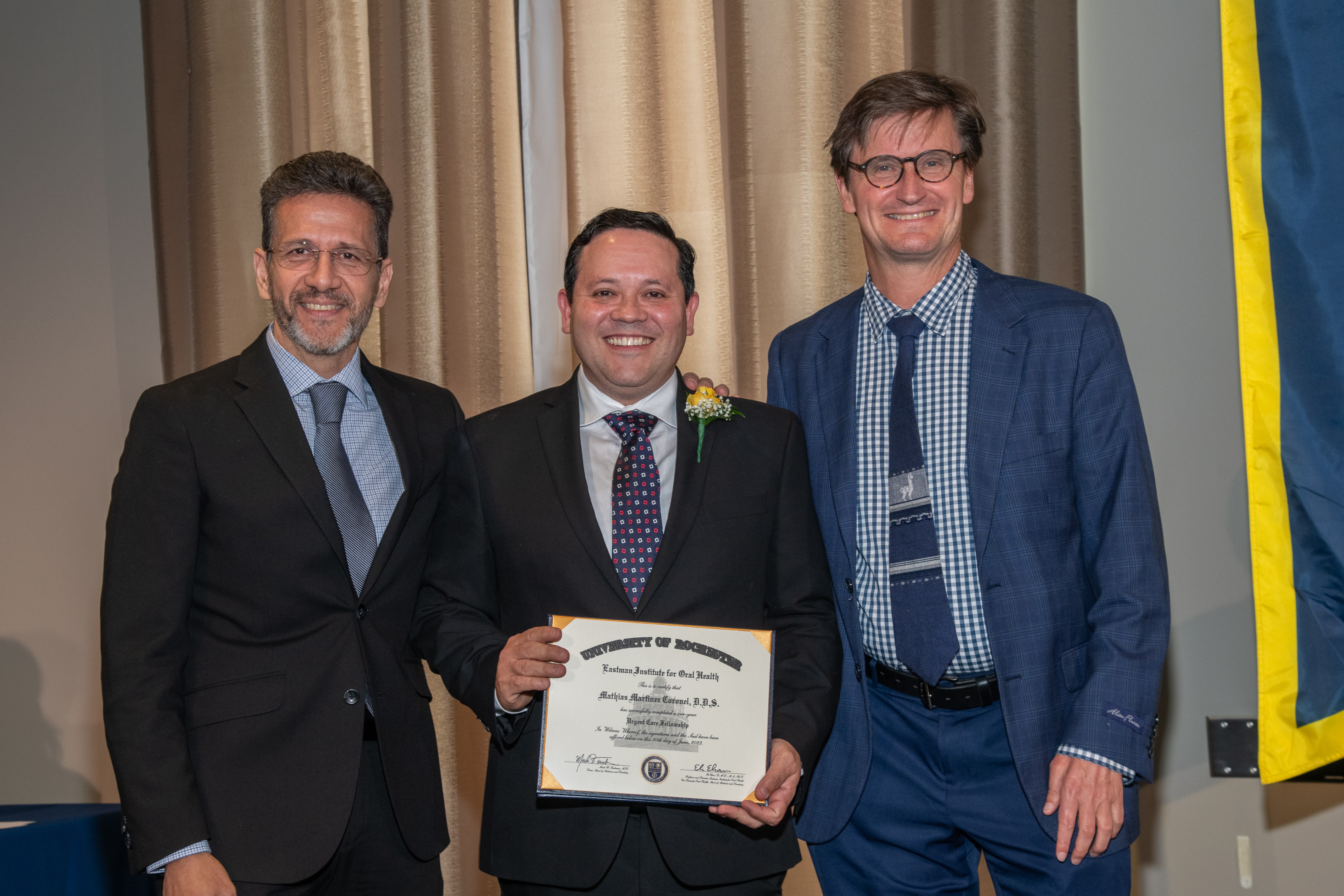3 men in suits. The man in the middle wears a boutonniere and holds a diploma.