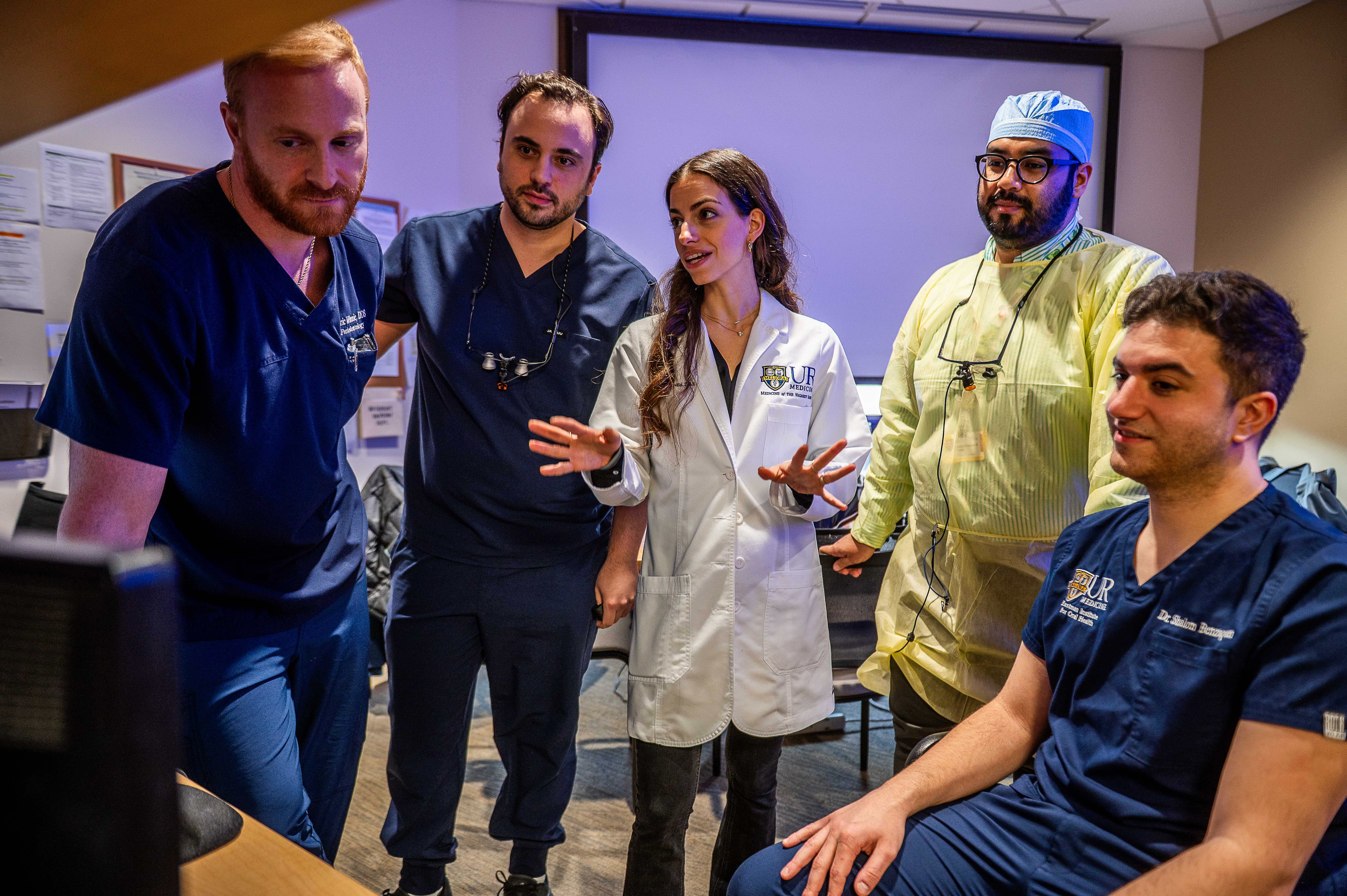 a woman in a lab coat explains something to 4 people in scrubs who are all looking at a computer screen.