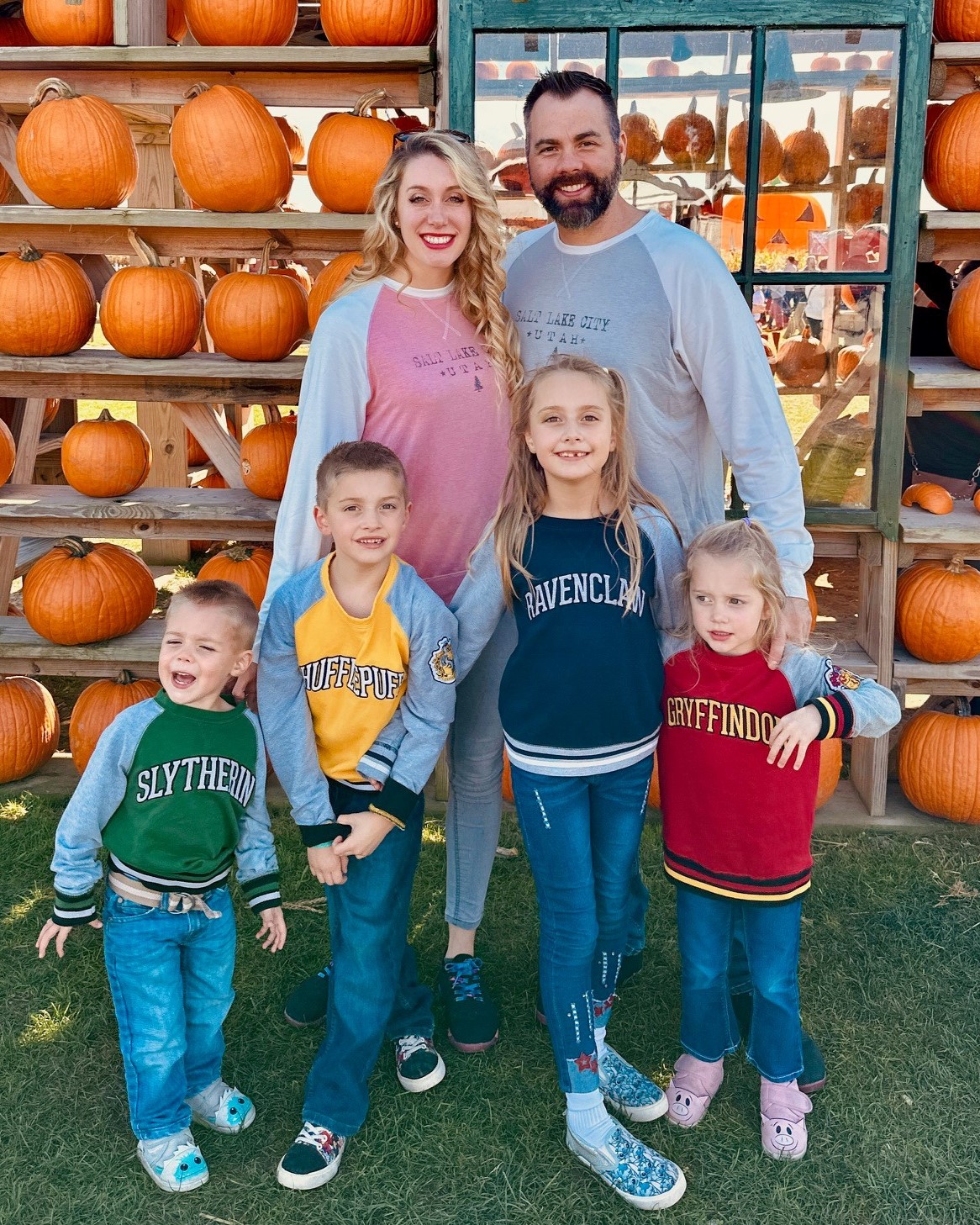 A man and woman stand behind 4 small children, who are wearing sweatshirts reading "Slytherin", "Hufflepuff", "Ravenclaw", and "Gryffindor". A shed filled with pumpkins is visible behind them.