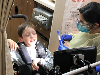 A patient in a wheelchair receives dental care on one of EIOH's SMILEMobiles a dentist in gloves, mask, and a yellow plastic gown stands next to a child in a wheelchair and holds a toothbrush and dental mirror.