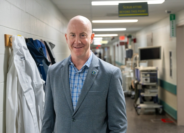 Ryan Fields stands in the hallway of the OR in Strong Memorial Hospital. Doctor coats, computers, and other equipment can be seen behind him. 