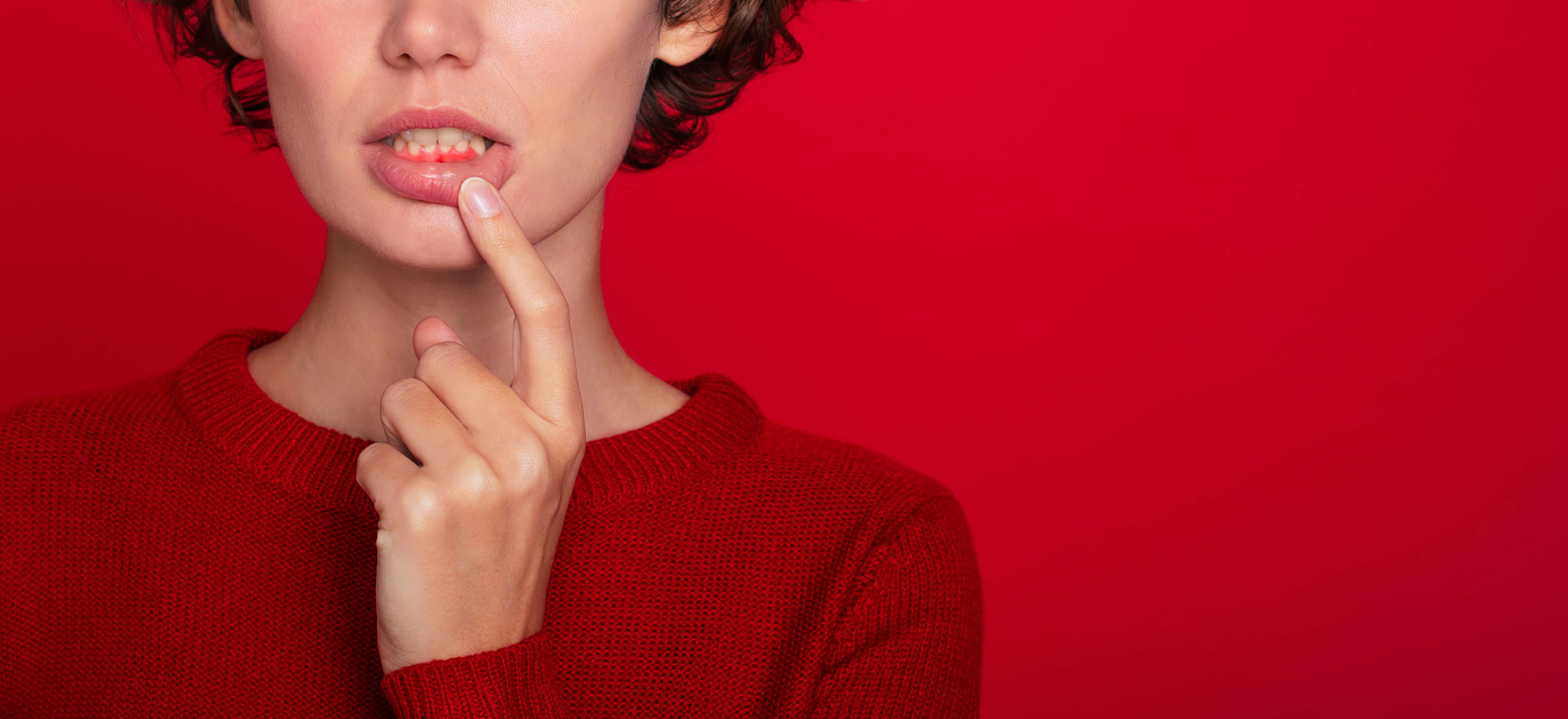 A woman points to her red gums, wearing a red top in front of a red background