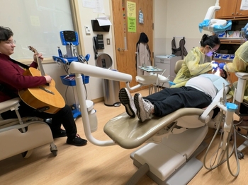 Two dental professionals work on the mouth of a pregnant patient while a woman plays guitar.
