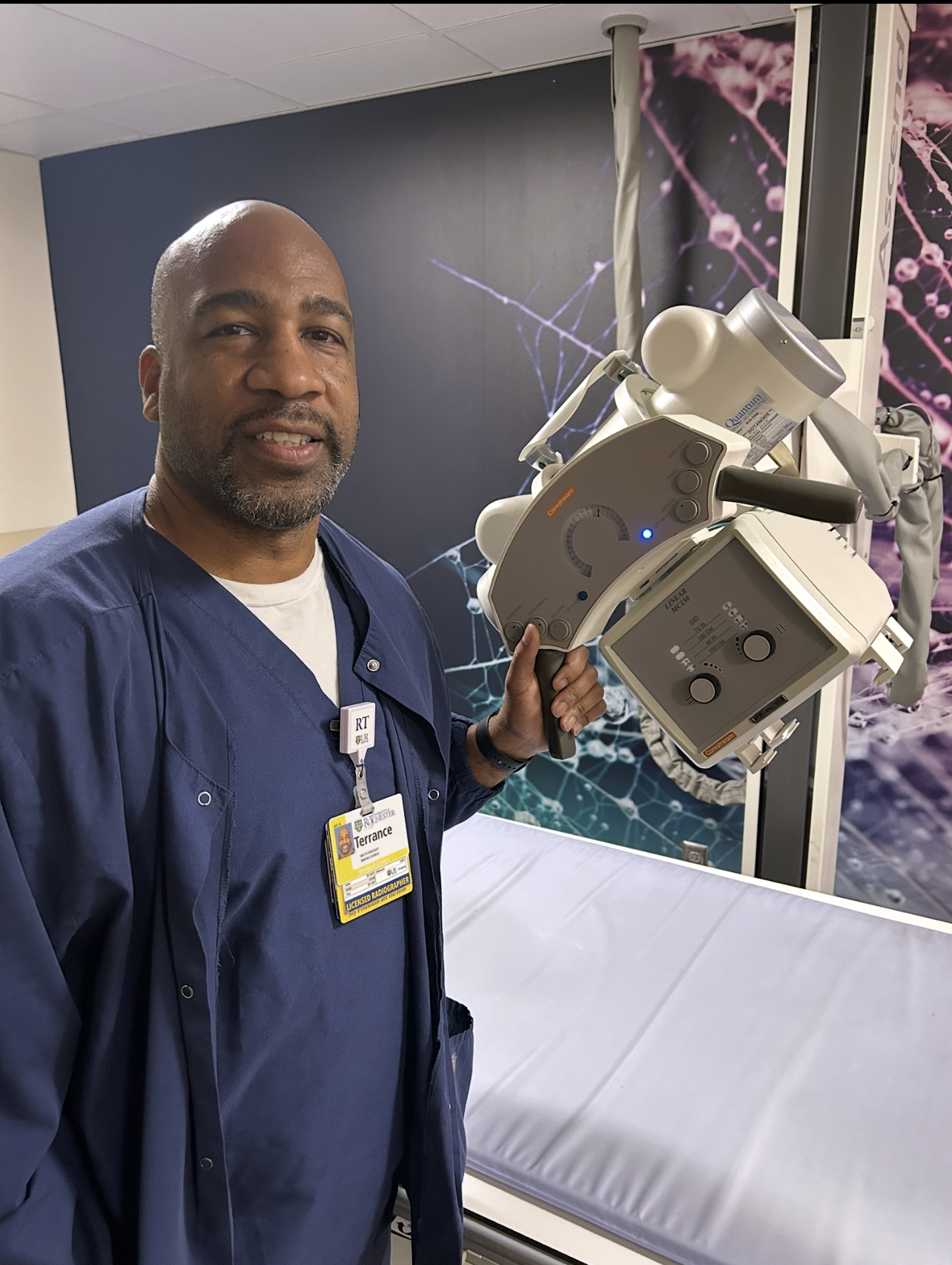 Terrance Cooper in blue scrubs operating an X-ray machine in a medical examination room. The scene includes a patient bed with a light gray mattress and a wall featuring a scientific, neuron-like design in purple and blue tones.