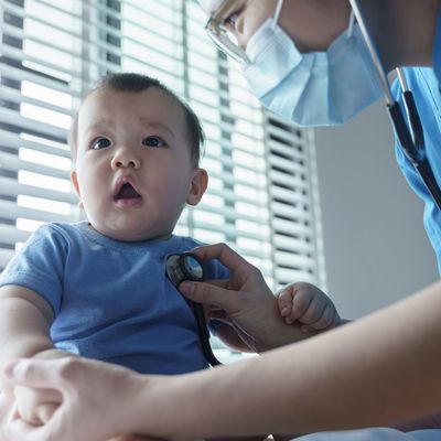 Asian Pediatrician in blue scrubs and a face mask using a stethoscope to examine a baby BOY in a blue shirt sitting on an examination table in a clinic.