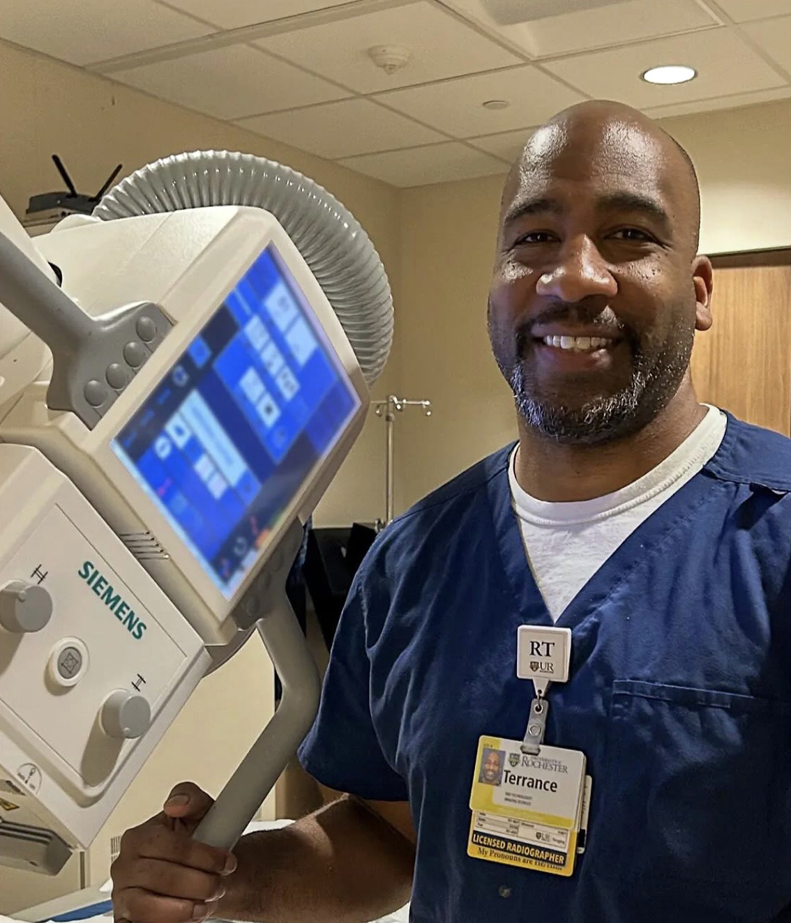Terrance Cooper in blue scrubs operating a Siemens radiography machine in a clinical setting. The scene highlights the his identification badge and the machine's control panel with blue and white interface elements.
