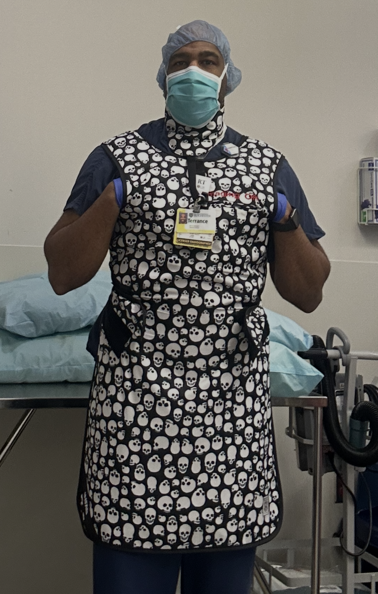 Terrance Cooper wearing a black and white skull-patterned protective apron and gloves, standing in a medical or clinical setting. He is giving two thumbs up, with medical equipment and a bed visible in the background.