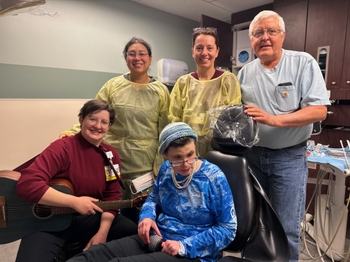 A female patient sits in the dental chair playing with an instrument called a cabasa. A woman with short hair sits next to her smiling and holding a guitar. A man in casual clothing and 2 dentists wearing  protective gowns stand behind smiling.
