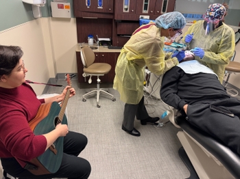 A patient of color lays calmly on the dental chair while two dental professionals use equipment in his mouth. A woman plays guitar nearby.