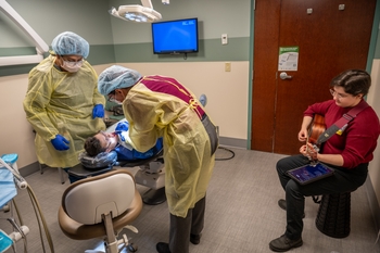 A patient lays in the dental chair while two dentists look in the patient's mouth. A woman plays guitar nearby.