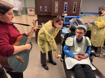 A female patient sits in the dental chair playing with a musical instrument while a dentist adjusts the chair. A woman plays guitar nearby.