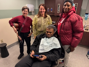 A patient of color smiles widely in the dental chair. Behind him stand his mother, as well as a woman holding a guitar and a dentist - all have big smiles.