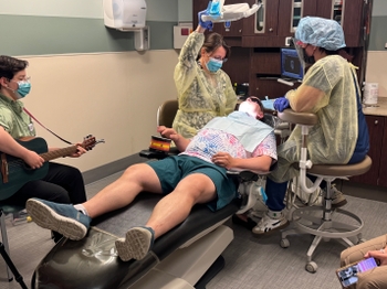 A male patient plays a small drum with one hand while he lays in the dental chair. Two dentists adjust a lamp to look in the patient's mouth. A woman plays guitar nearby.