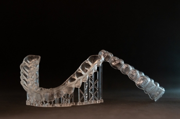Two clear orthodontic aligners in front of a black background. One has been 3D-printed and is still connected to a plastic scaffold..