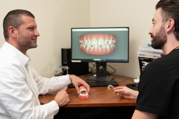 A man wearing a white lab coat talks with a man wearing black scrubs. They hold a clear orthodontic aligner and a physical model of a mouth, and sit in front of a computer screen displaying a digital version of the model. 
