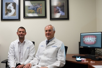 Two men wearing white lab coats sit in front of a computer screen displaying a 3D model of a mouth.