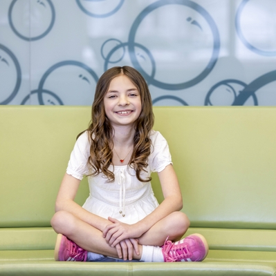 young girl in white dress sitting cross legged on couch smiling