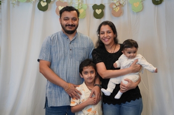 A young man and woman smile in front of a white backdrop and decorations shaped like baby clothes. The man has his arms around a young boy, and the woman holds a curious infant. 