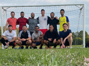 Twelve men pose together in front of a soccer goal, wearing athletic clothing. The six in the back stand with their arms around each others' shoulders, while the other six squat in front of them.