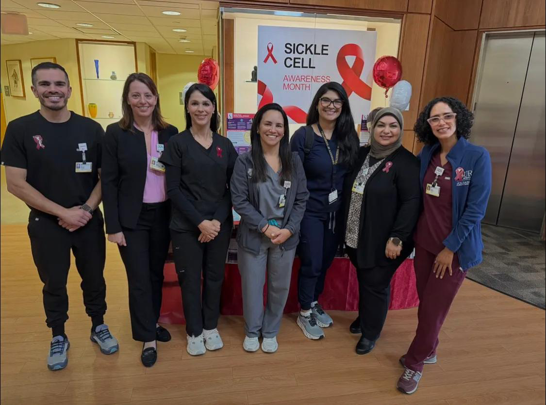 Seven people wearing scrubs stand in front of a table with informational papers on it, in the Eastman Dental Atrium. Behind them is a "Sickle Cell Awareness Month" poster, and red and white balloons.