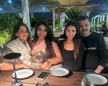 Three women and a man sit together at an outdoor table, with their arms around each other. One of the women has a small fluffy dog on her lap. 