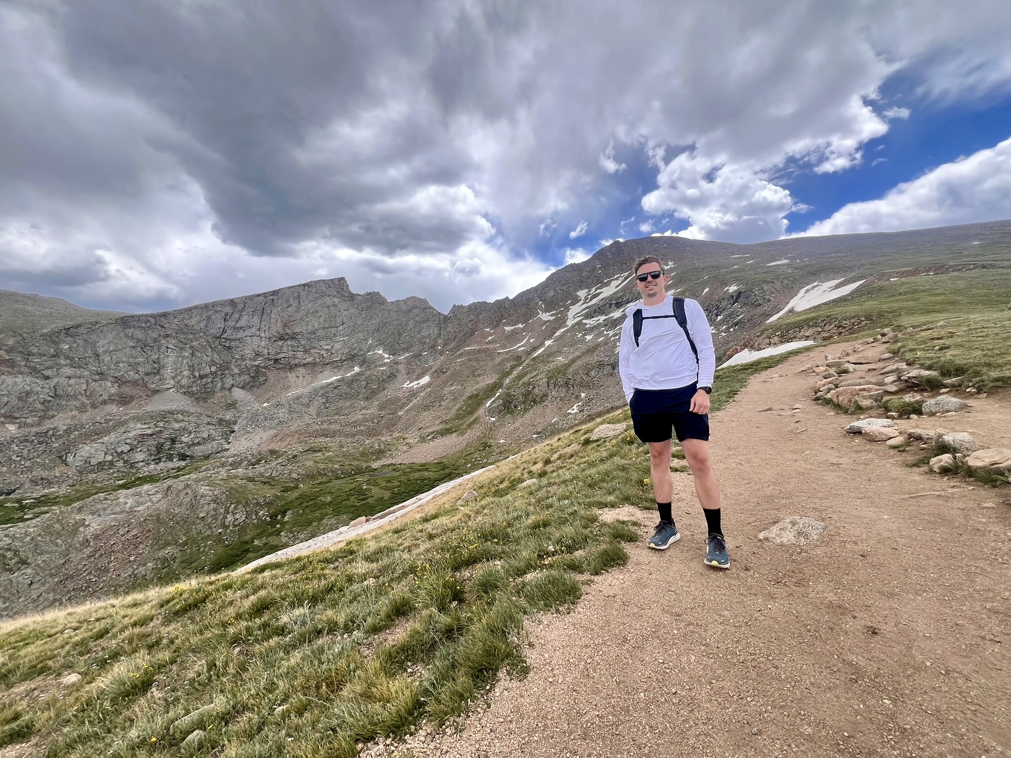 A man stands on a dirt path that curves up a rocky, scrub-covered mountain. The sky above is a deep blue filled with thick clouds.
