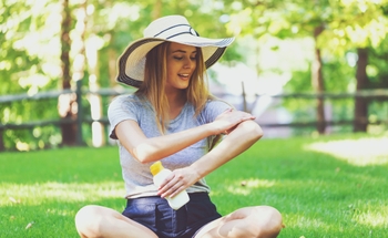 Woman sitting outdoors applies sunscreen and wears hat