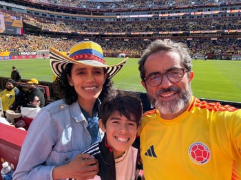 A woman with dark curly hair wearing a colorful hat hugs a boy and stands close to a bearded man wearing a yellow soccer jersey. They are in the bleachers of a soccer stadium.
