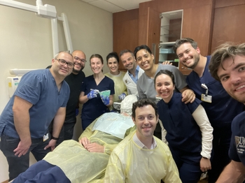A large group of dentists wearing scrubs or lab coats cluster around a dental chair, smiling. One of them is sitting in the dental chair, face completely covered in a white plaster-like substance except for a straw that pokes out of their mouth.