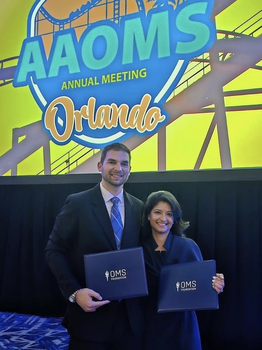 A tall man and a shorter woman hold up certificate covers that say "OMS Foundation". Behind them is a brightly colored screen announcing the AAOMS Annual Meeting in Orlando.