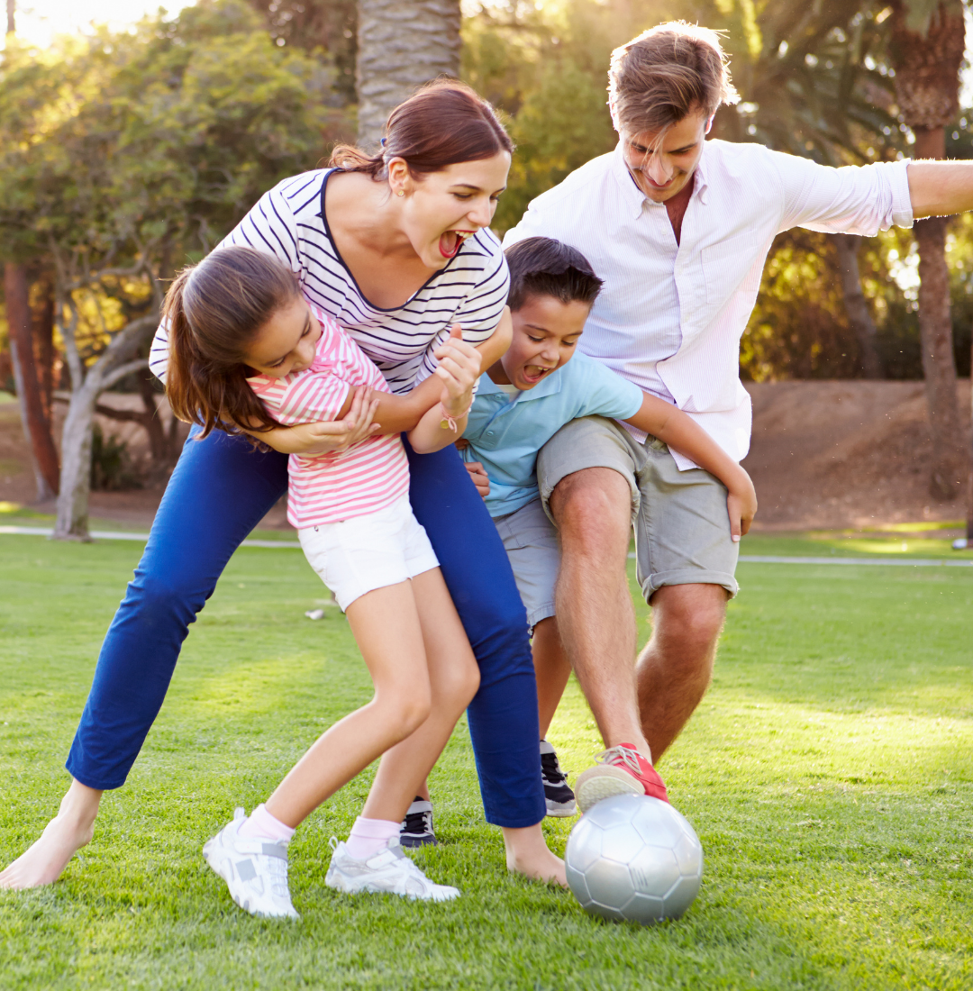 A family plays soccer on a grassy field with palm trees in the background.