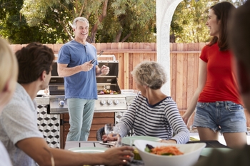 A man stands at the grill filled with veggie kababs while a group surrounds him smiling. 
