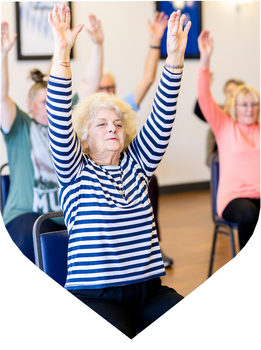 Older adults participate in a seated exercise class, raising their arms overhead during a group fitness session.