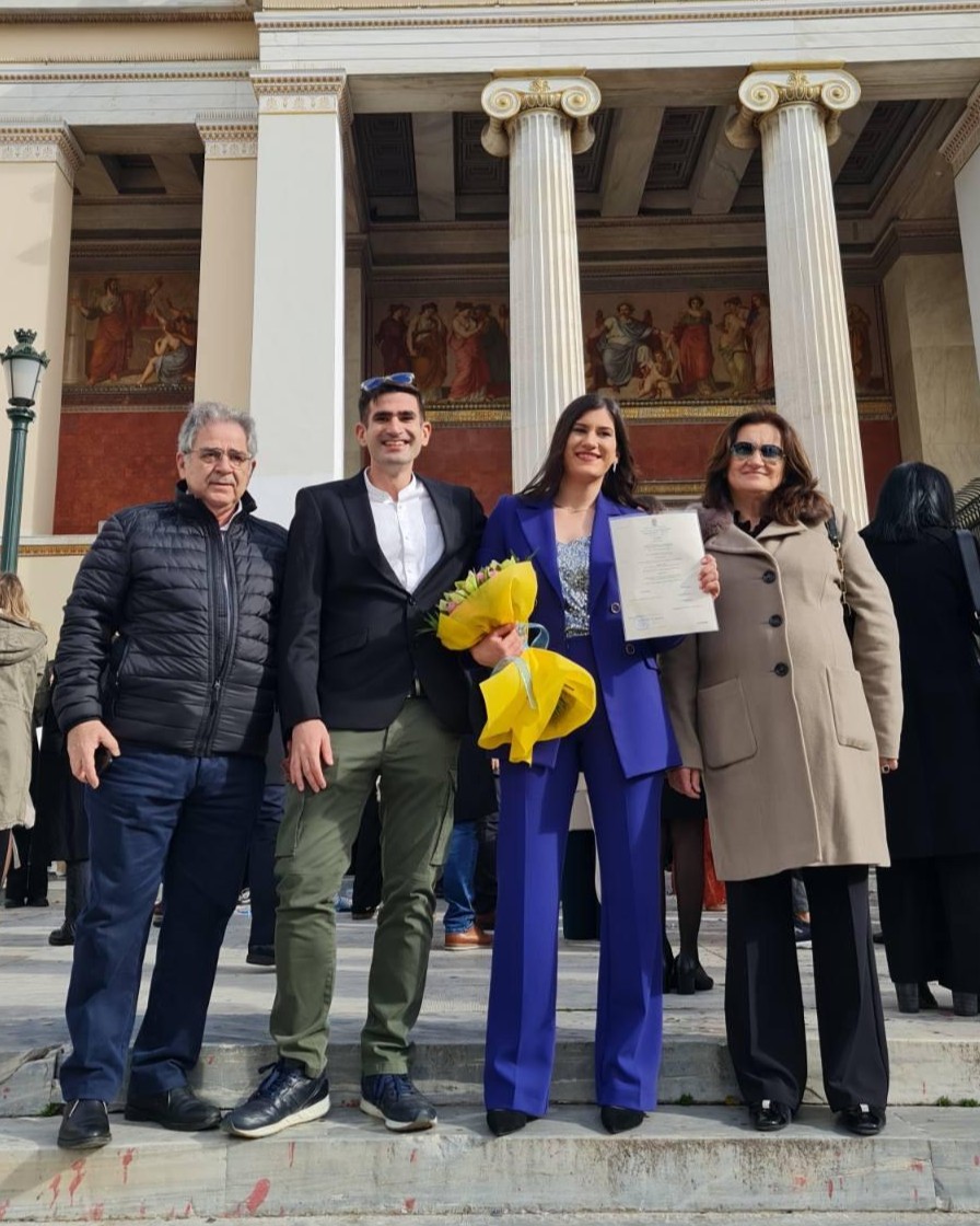 A young woman smiles on the steps of a columned building, holding a bouquet of flowers and a certificate. Beside her are a young man, as well as an older man and woman.
