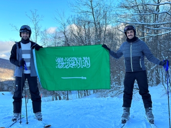 Two young men stand on skis at the top of a snowy slope, holding a large Saudi Arabia flag between them.