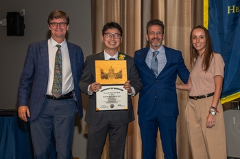 Three men and a woman wearing formal attire pose together at a graduation. The man in the center holds a certificate.