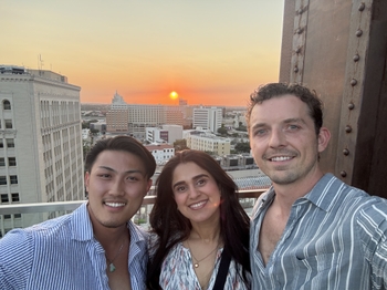 Two men and a woman stand on a balcony overlooking downtown Rochester, NY. Behind the tall buildings is a soft orange sunset that fades into a white sky overhead.