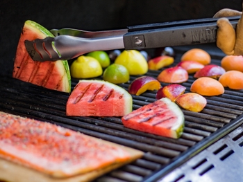 A selection of fruit is seen on a grill including a slice of watermelon between grill tongs and peaches in the background.