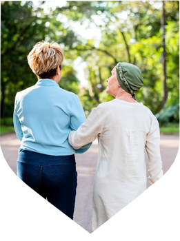 Two women walk arm-in-arm outdoors on a tree-lined path. One woman wears a head covering commonly worn during cancer treatment.